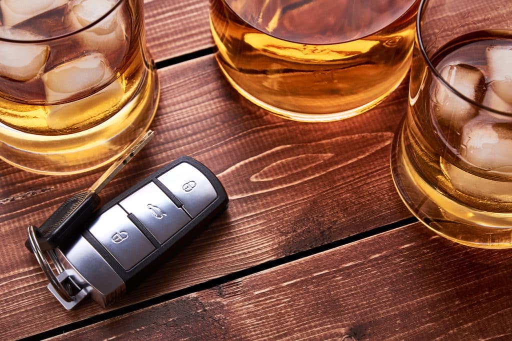 Still life on old wooden table top. Car keys, several glasses and a bottle of whiskey or alcohol. Suitable for drunk driving.