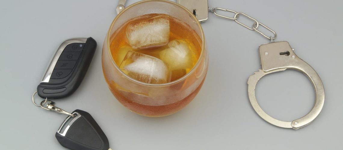 Glass with alcohol, steel handcuffs and car key on gray background.